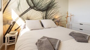 A bedroom with a large bed, white bedding, a nightstand with a lamp, and a wall mural. - Au Gré du Vent by Interhome (Brignogan-Plage)