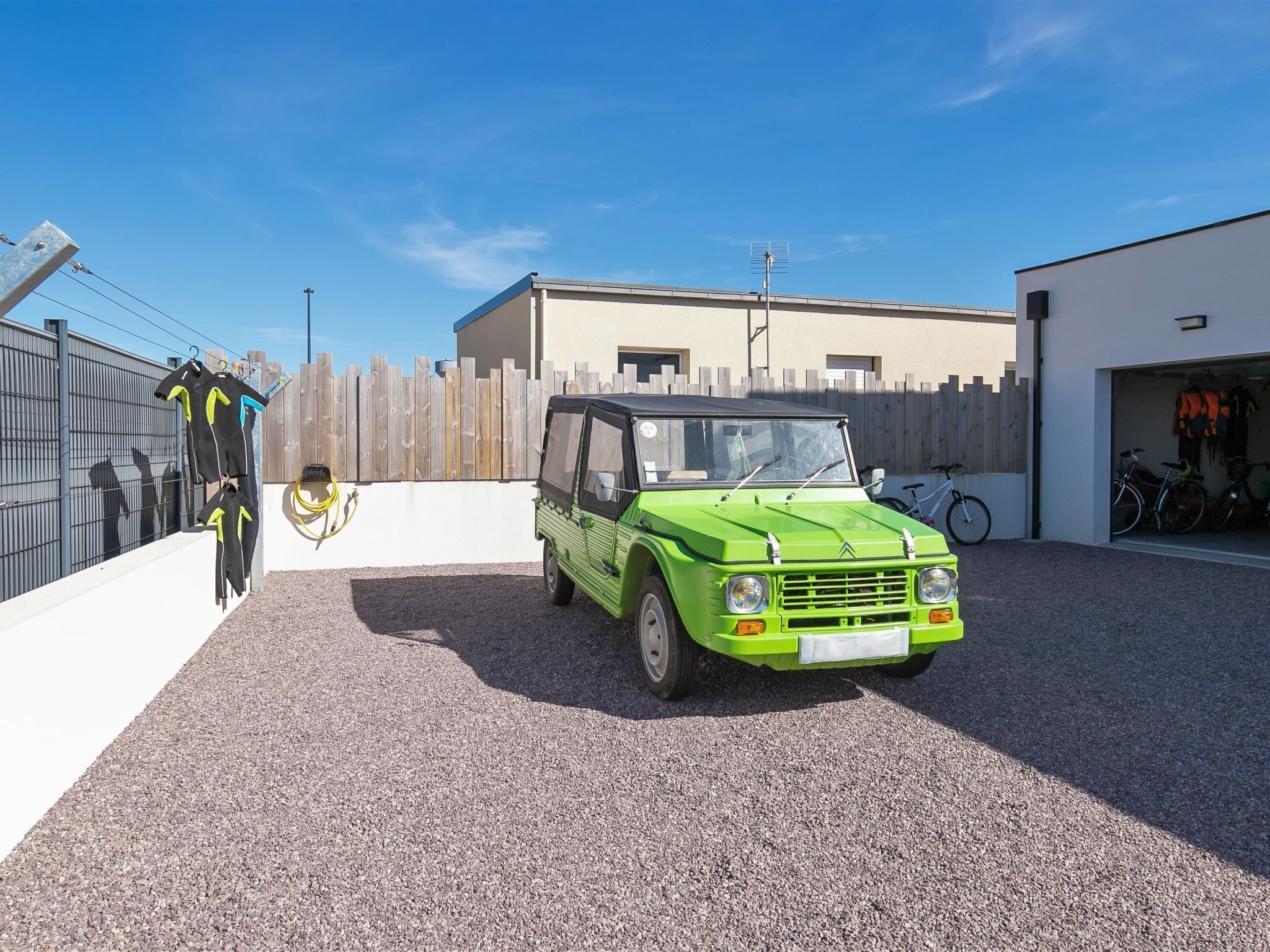 Un coche verde aparcado en un camino de grava con una valla de madera y un edificio al fondo.
