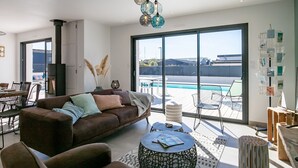 A modern living room with a brown sofa, a coffee table, and a view of a swimming pool through large windows. - Au Gré du Vent by Interhome (Brignogan-Plage)