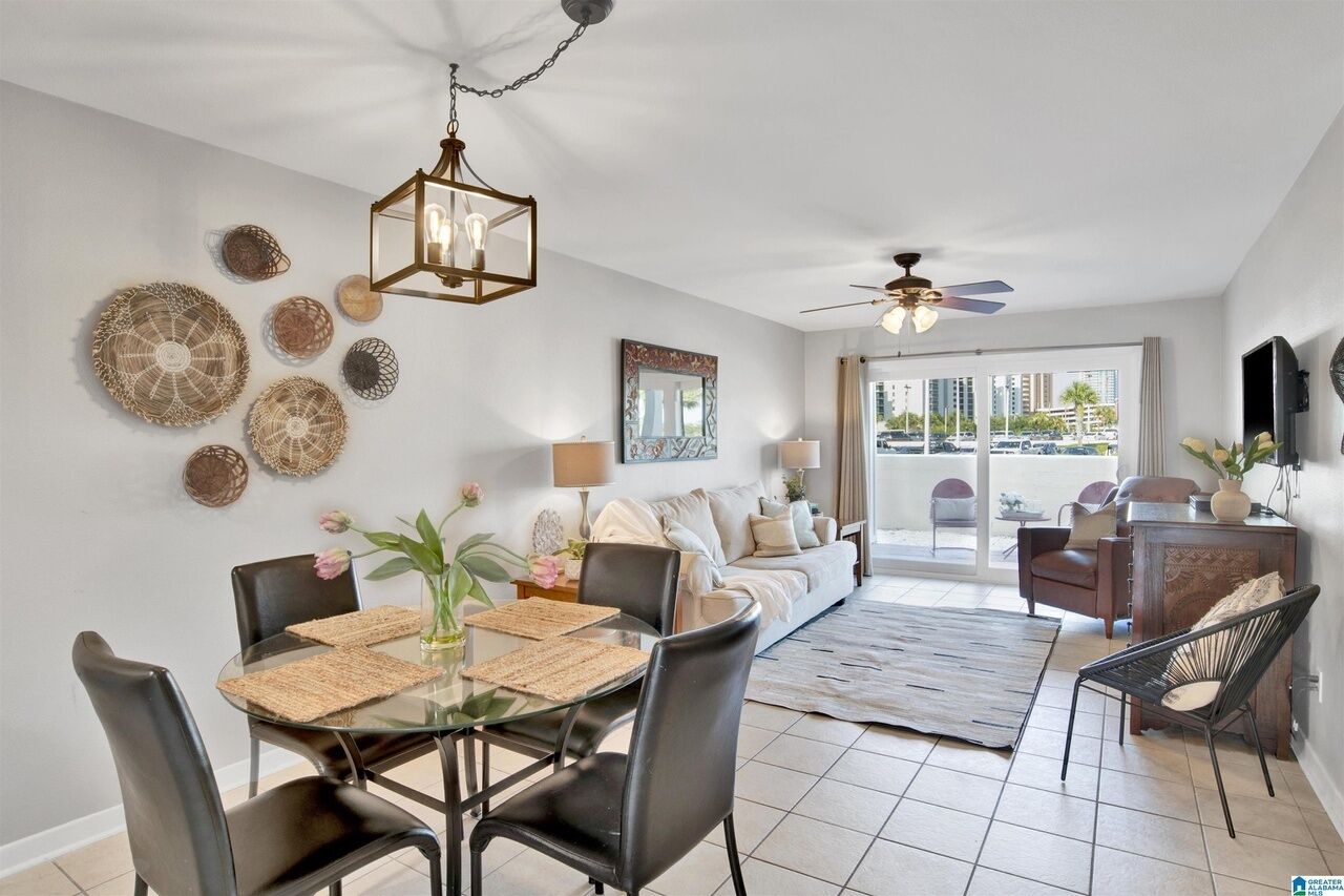 A modern living room with a dining area, featuring a ceiling fan, a chandelier, and a large window with a view of the city.