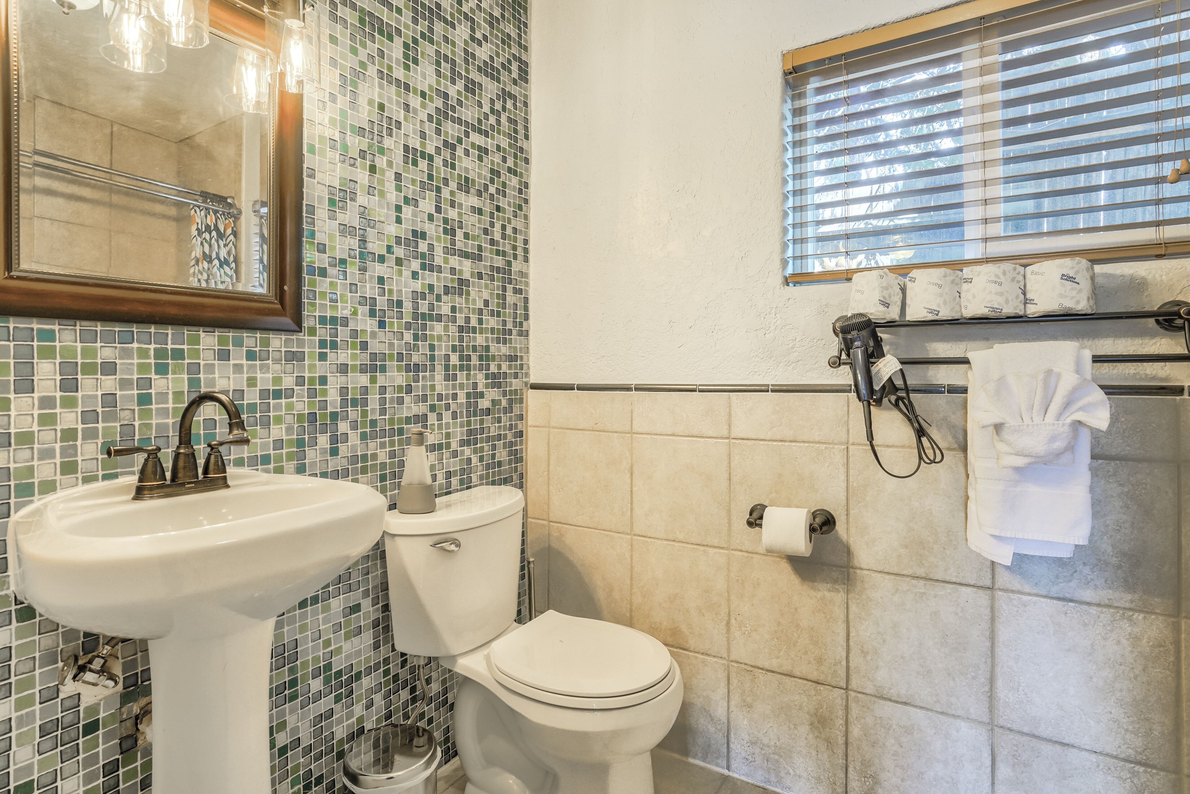 A bathroom with a mosaic tile wall, a white sink, a toilet, and a window with blinds.