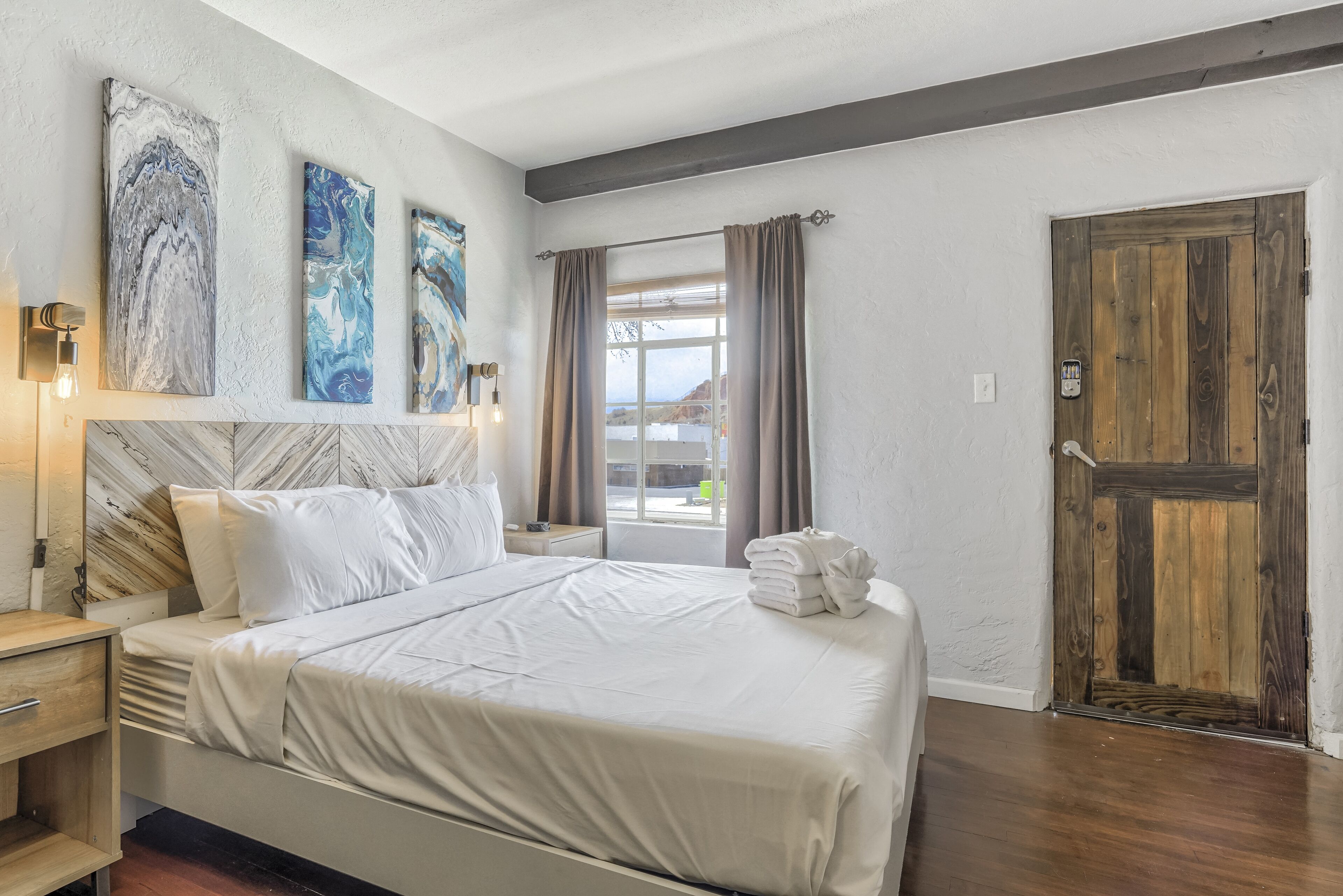 A neatly made bed with white linens, a wooden headboard, and a nightstand with a lamp. There are three abstract paintings on the wall, and a wooden door is visible in the background.