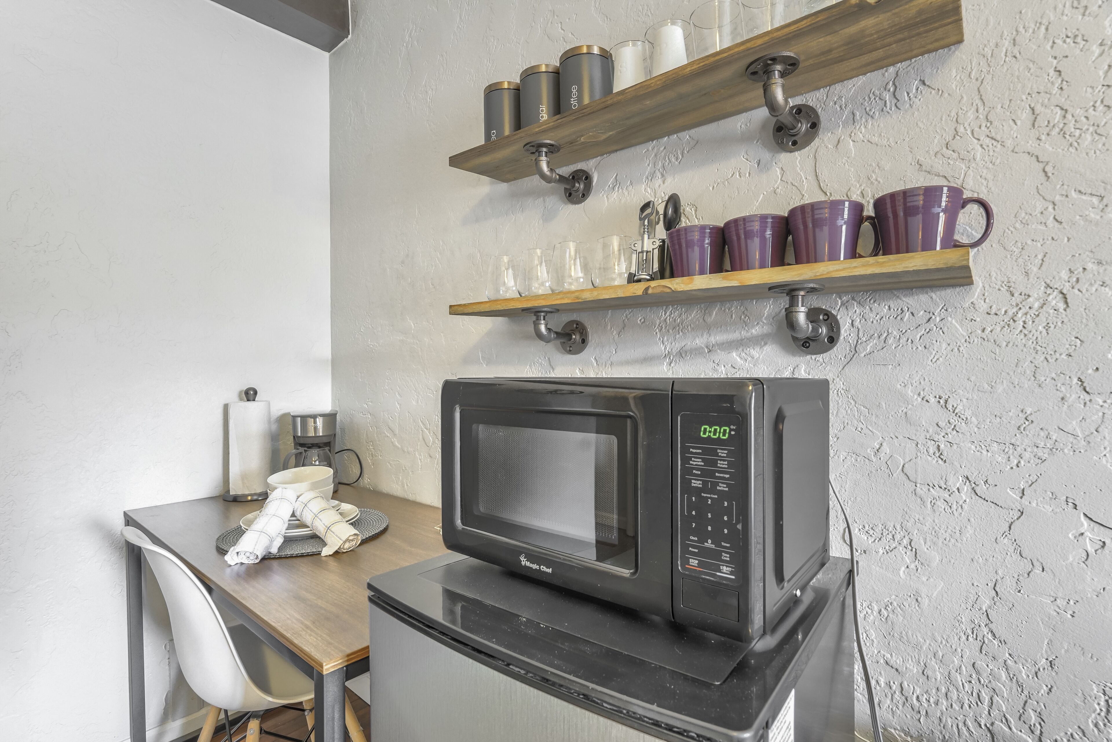 A compact kitchen with a microwave on a counter, a small table with a chair, and shelves above holding mugs and glasses.