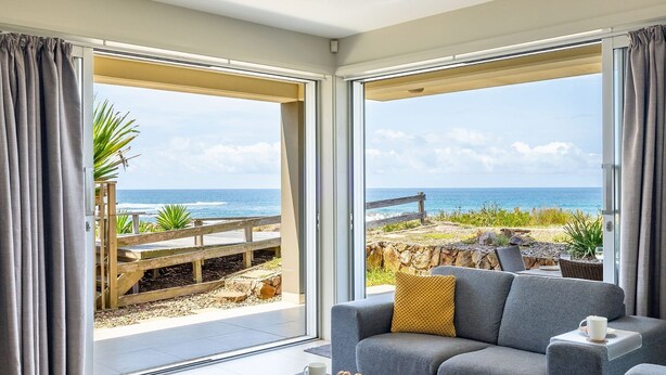 A room with a grey sofa, a wooden deck, and a view of the ocean. - Ocean on the Rocks, 1,21 Ocean Ave - waterfront, Wi-Fi, Ducted Air Conditioning (Anna Bay)