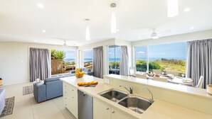 A modern kitchen with a large window overlooking the beach, a dining area with a table set for two, and a cozy seating area with a sofa. - Ocean on the Rocks, 1,21 Ocean Ave - waterfront, Wi-Fi, Ducted Air Conditioning (Anna Bay)