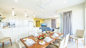 A modern dining area with a wooden table set for a meal, white chairs, a kitchen area with a microwave, and a TV in the background. - Ocean on the Rocks, 1,21 Ocean Ave - waterfront, Wi-Fi, Ducted Air Conditioning (Anna Bay)