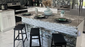 A modern kitchen with white cabinets, a stainless steel refrigerator, a microwave, and a coffee maker. There are three black stools at the island, and a vase with dried plants.