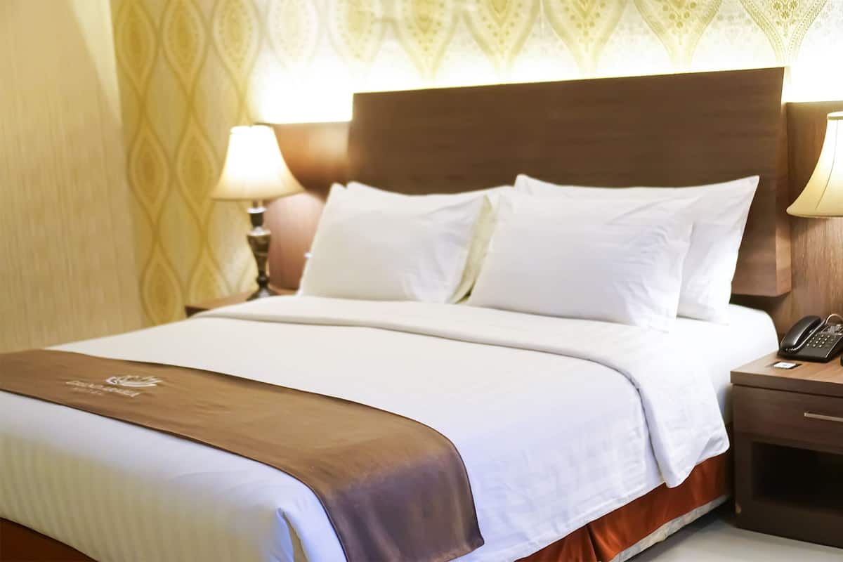 Una cama bien hecha con sábanas blancas y detalles en marrón, una lámpara de noche y un teléfono sobre una mesita de madera.