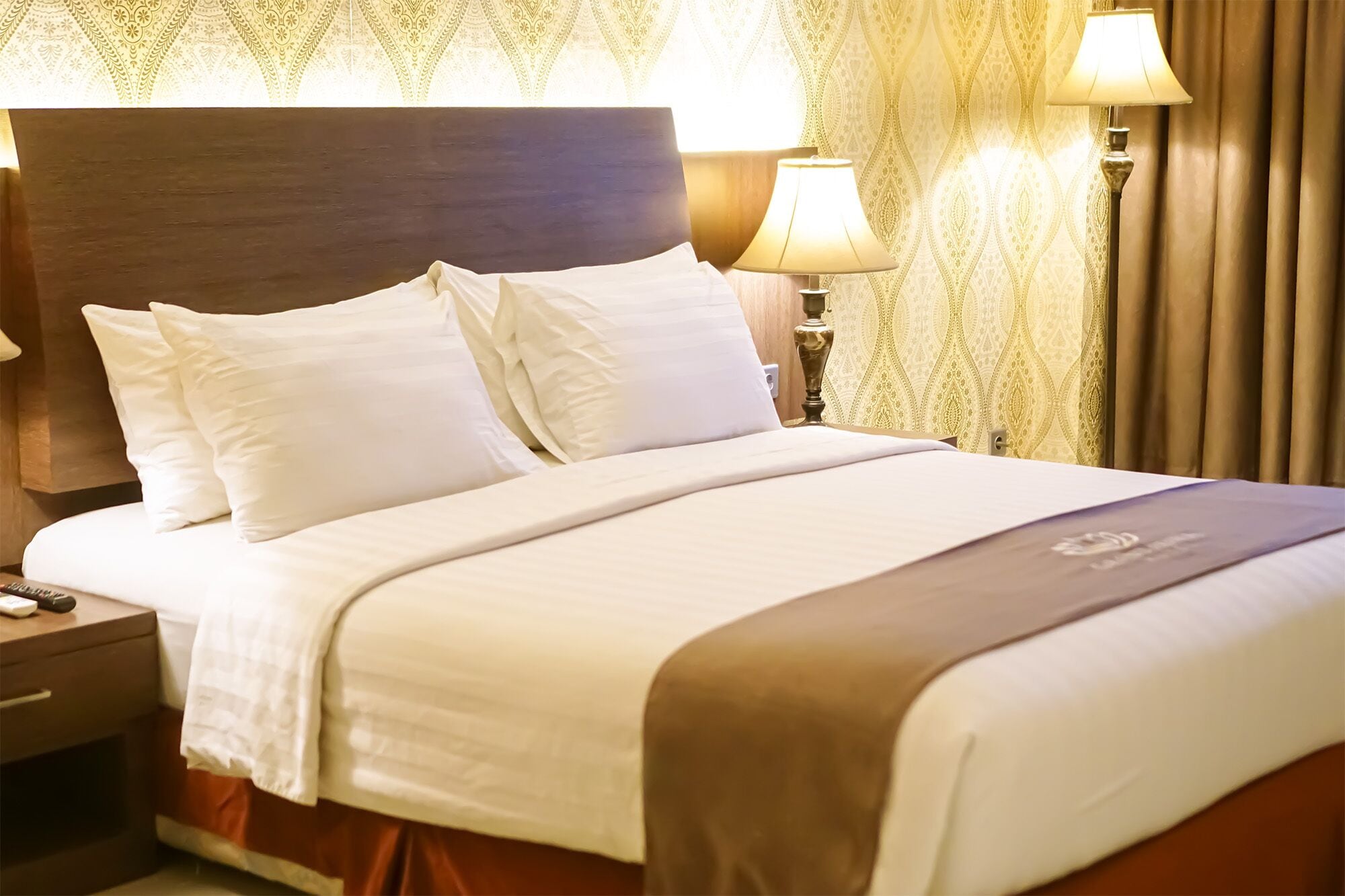 A neatly made bed with white linens and a brown accent throw, a wooden nightstand with a lamp, and patterned curtains.