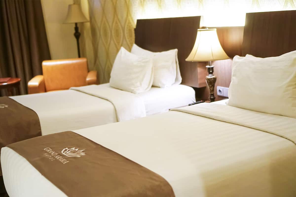 Habitación de hotel con dos camas, cada una con ropa blanca y cabeceros marrones. Hay una lámpara en la mesita de noche y una silla en la esquina.