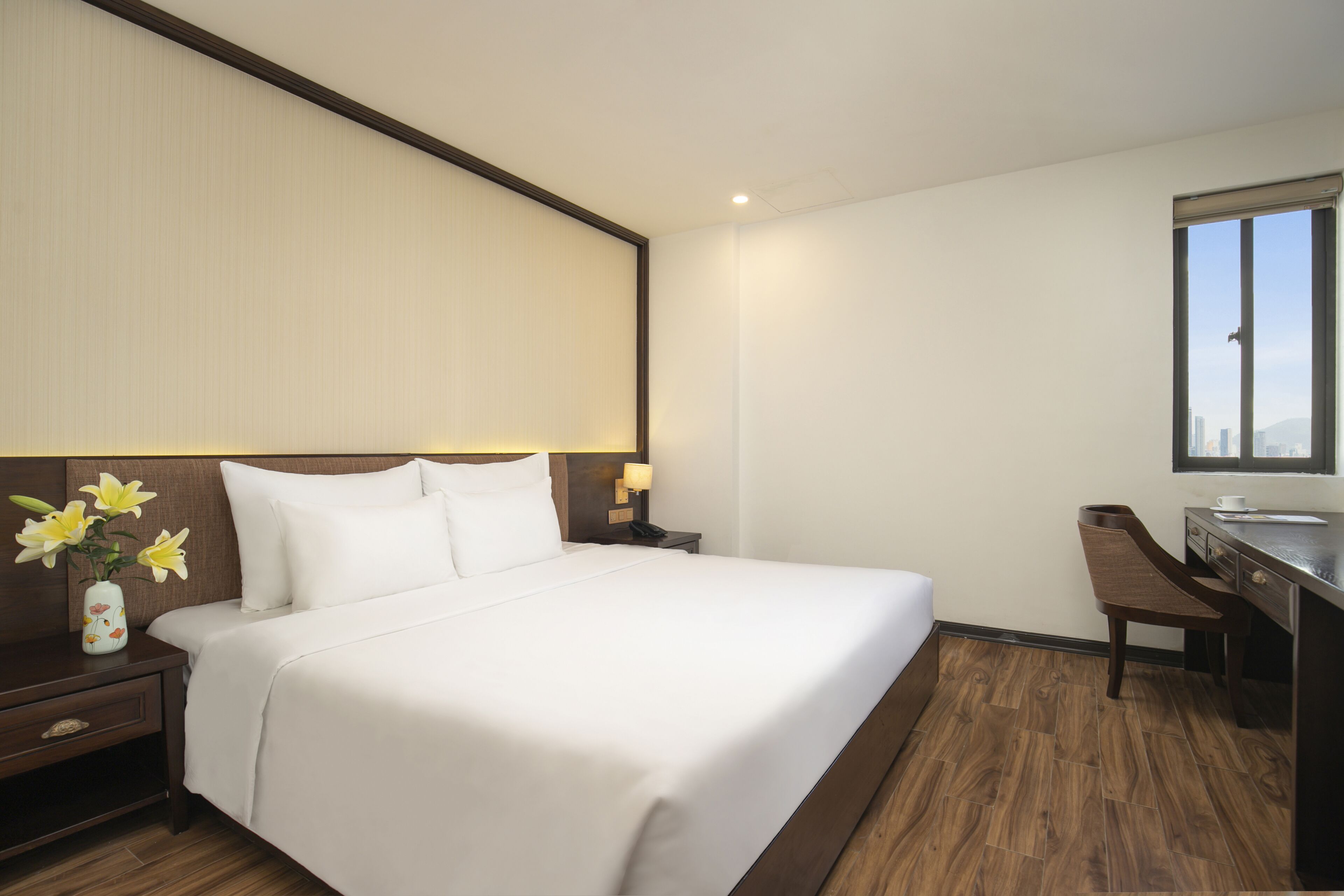 A hotel room with a large bed, a nightstand with a vase of flowers, a desk with a chair, and a window with a view.