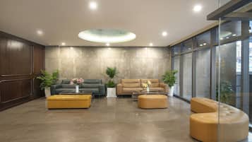 A modern lobby with two sofas, a yellow ottoman, and potted plants.