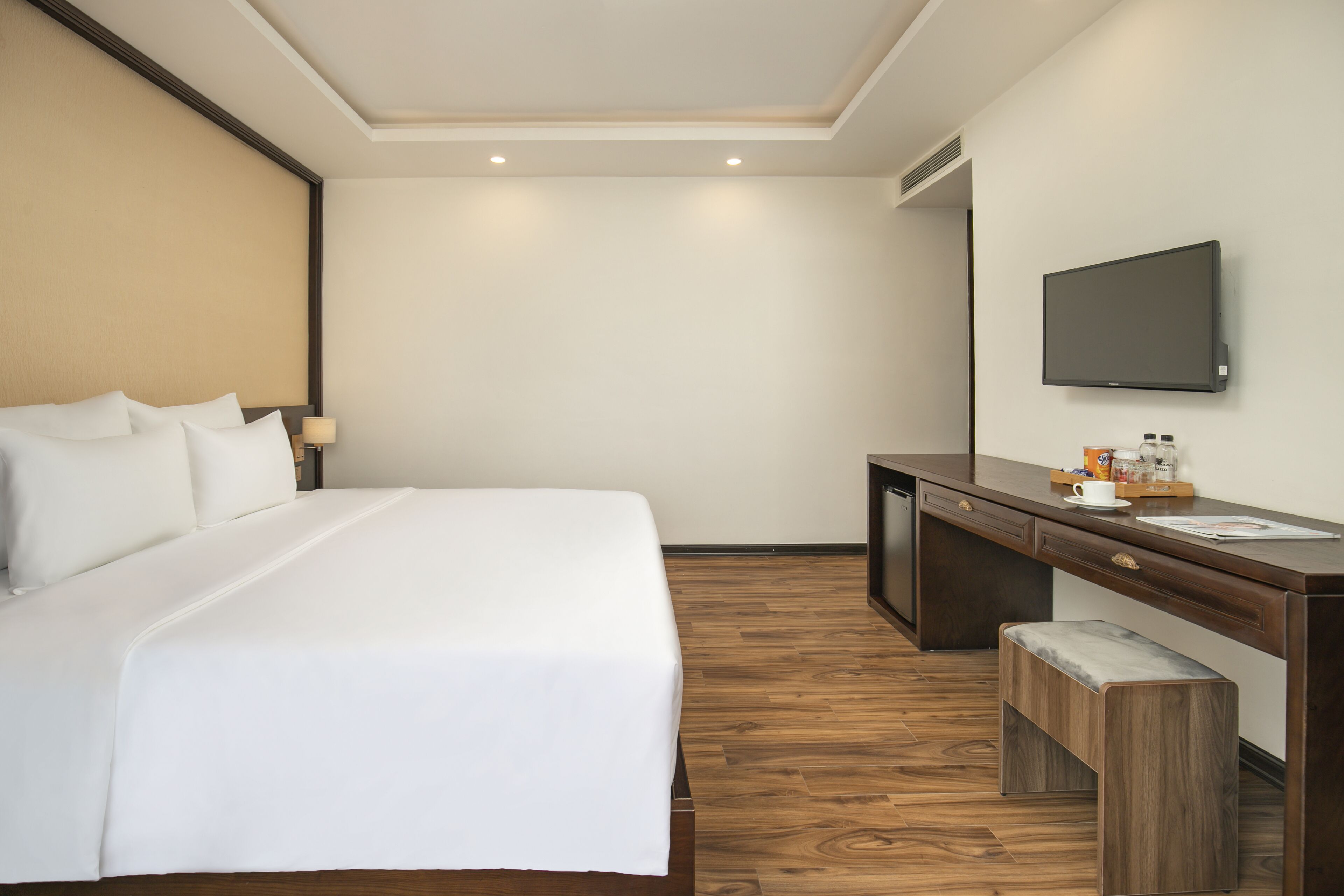 A hotel room with a large bed, a desk with a mirror, a television, and a small bench.