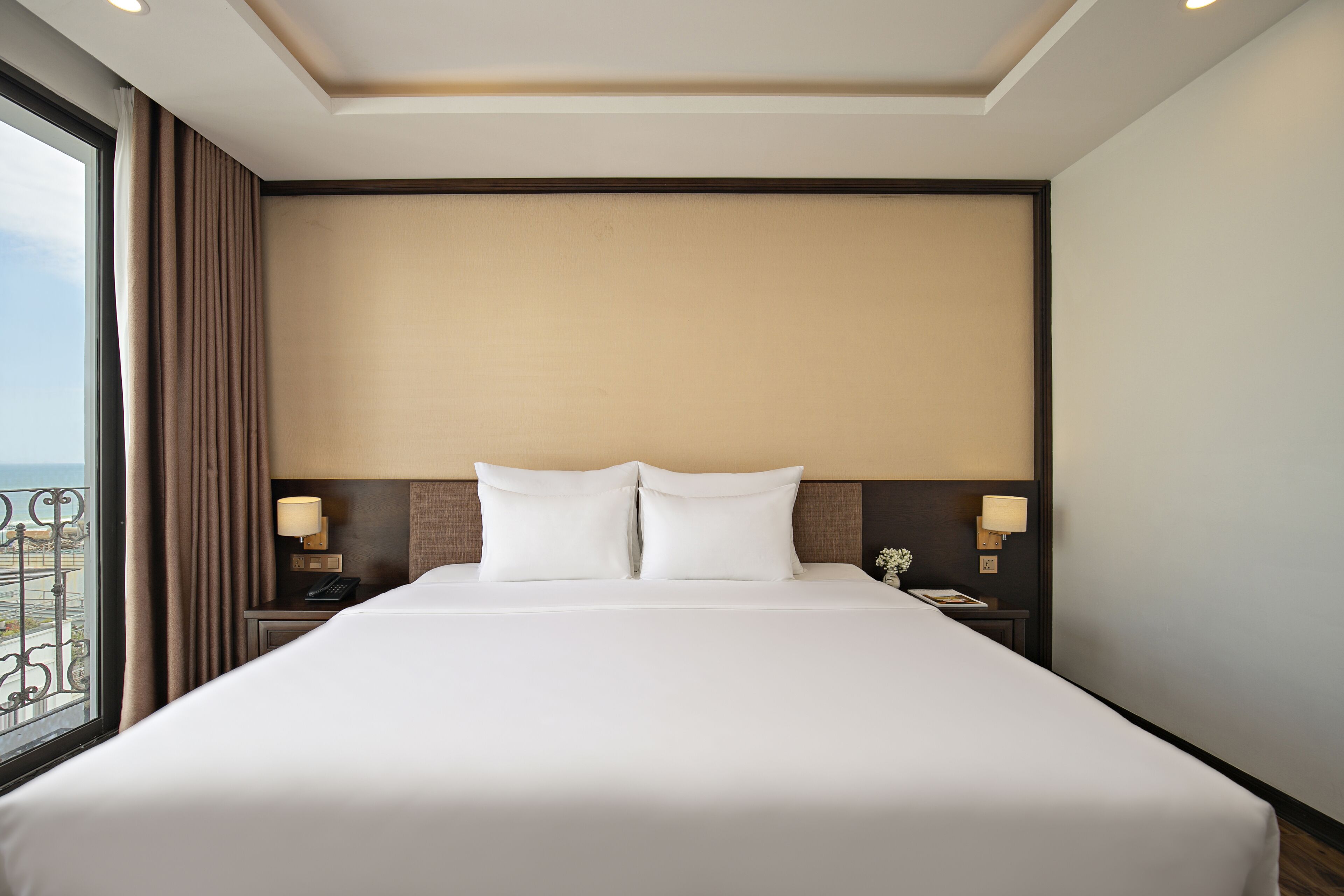 Habitación de hotel con una cama grande, dos mesitas de noche con lámparas, vistas al exterior a través de una ventana y un cabecero de color neutro.