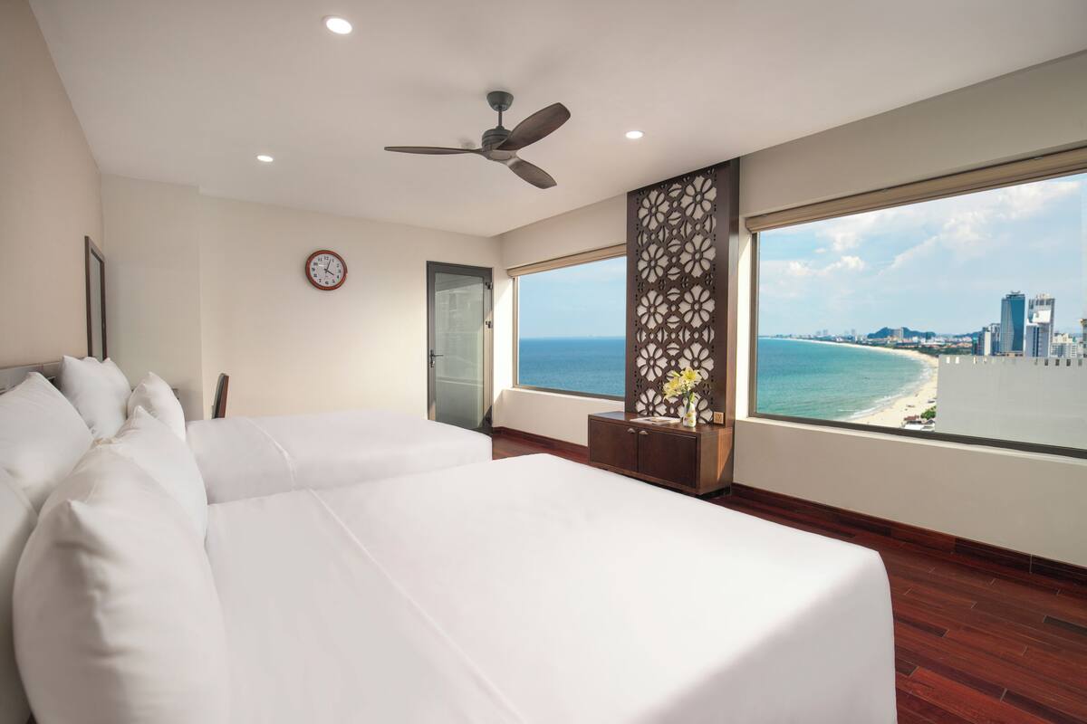 Ein Hotelzimmer mit einem großen Bett, einem Deckenventilator, einer Wanduhr, einem Fenster mit Blick auf Stadt und Strand sowie einer dekorativen Wandverkleidung.
