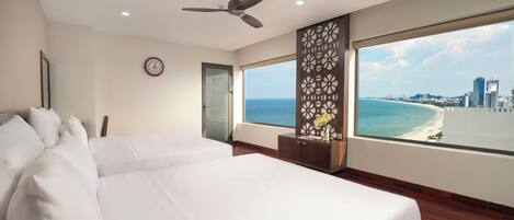 A hotel room with a large bed, a ceiling fan, a clock on the wall, a window with a city and beach view, and a decorative panel.