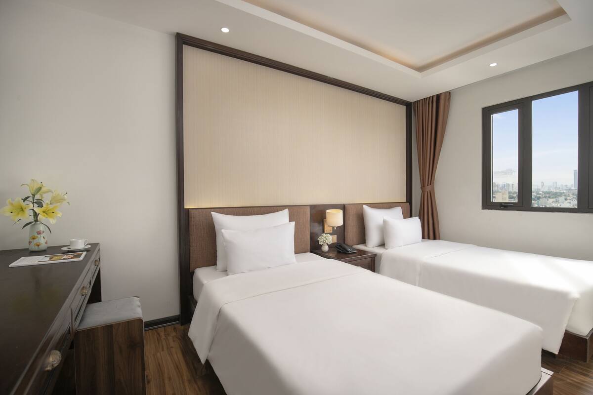 Ein Hotelzimmer mit zwei Betten, einem Schreibtisch, einem Nachttisch, einer Lampe und einem Fenster mit Blick auf die Stadt.