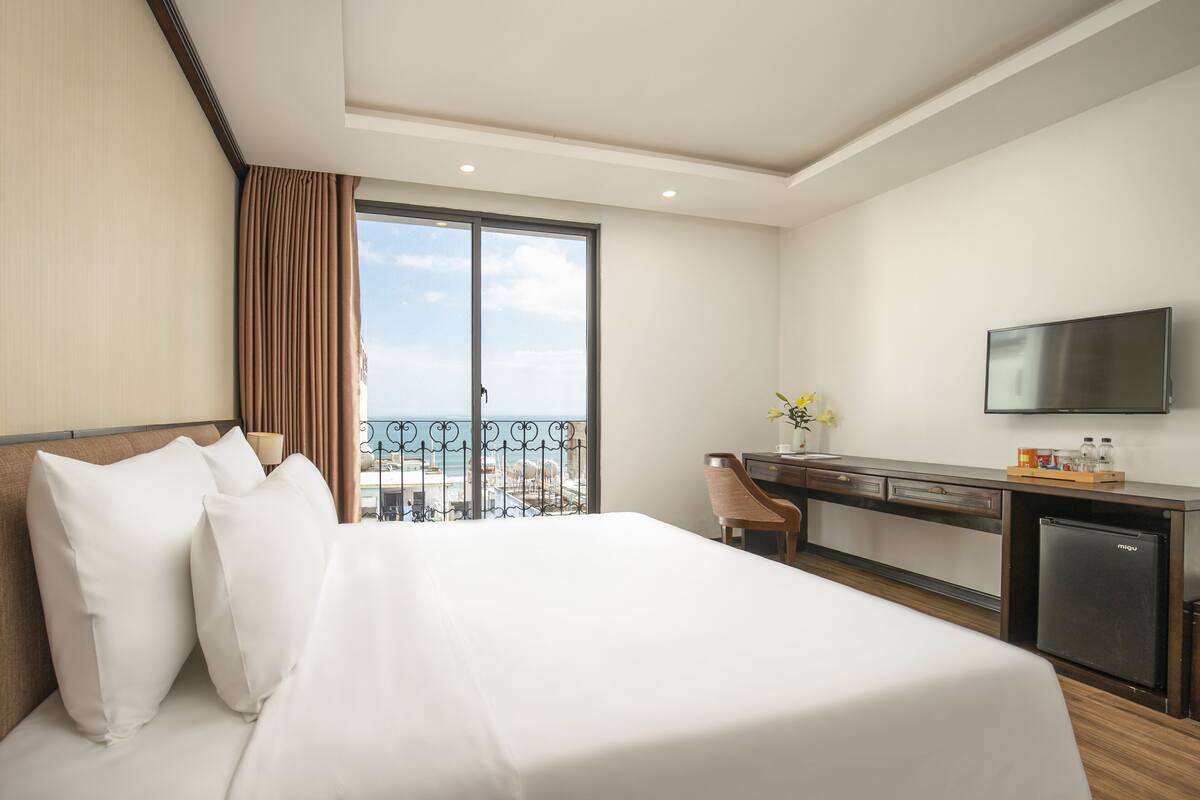 Ein Hotelzimmer mit einem großen Bett, einem Schreibtisch mit Computer, einem Fernseher und einem Balkon mit Meerblick.
