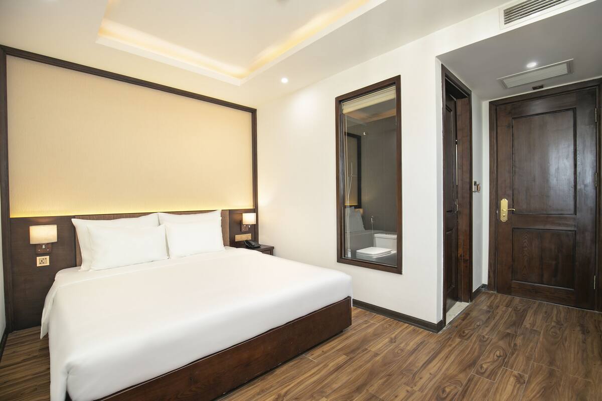 Ein Hotelzimmer mit einem großen Bett, einem Spiegel, einer Holztür und einem Nachttisch mit einer Lampe.