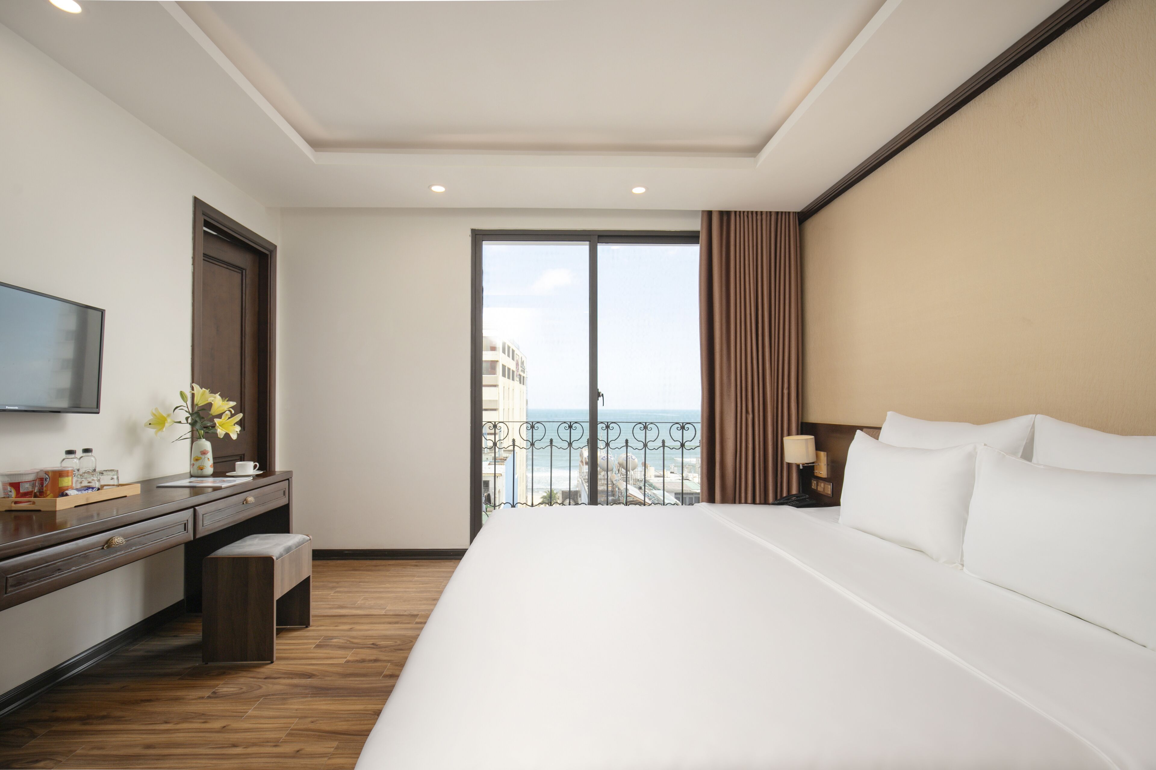 Quarto de hotel com uma cama grande, uma escrivaninha com TV de tela plana, uma mesinha com um vaso de flores e uma varanda com vista para a cidade.