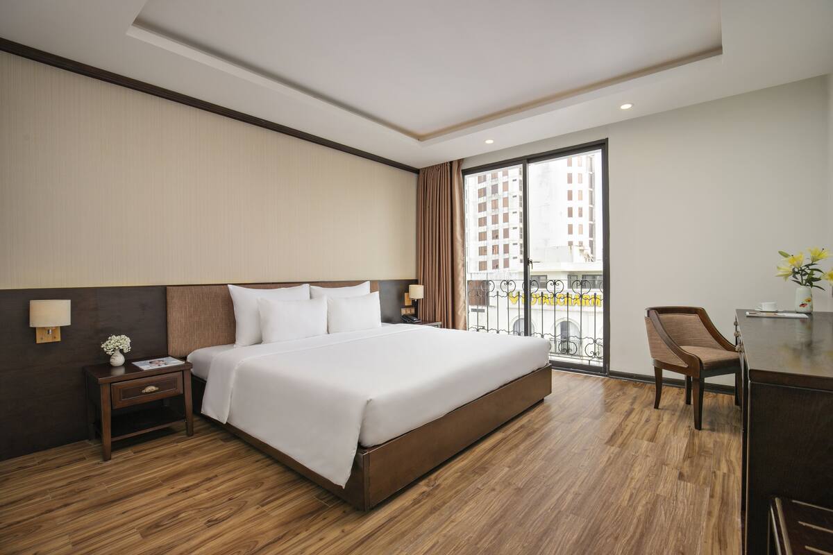 Ein Hotelzimmer mit einem großen Bett, einem Schreibtisch mit Stuhl, einem Nachttisch und einem Fenster mit Blick auf Gebäude.