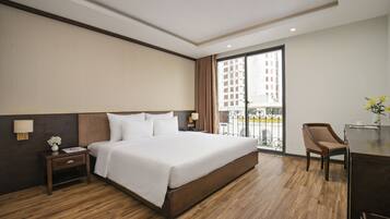 A hotel room with a large bed, a desk with a chair, a nightstand, and a window with a view of buildings.