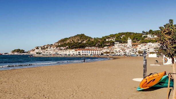 A beach with a clear view of the sea, a sandy shore, and a town in the background with buildings and a hill.