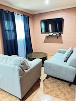 Un salón con dos sofás grises, una mesa de centro redonda, un televisor de pantalla plana montado en una pared de ladrillo y una ventana con cortinas azules.