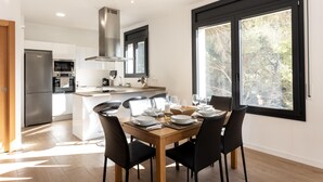 Une cuisine moderne dotée d’un îlot central, d’un coin repas avec une table en bois et des chaises noires, et de grandes fenêtres offrant une vue sur les arbres.