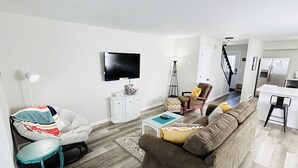 A modern living room with a sofa, armchair, and a coffee table. A television is mounted on the wall, and a kitchen area is visible in the background. - Highland Hideaway-Newly Remodeled!  (Sheridan)