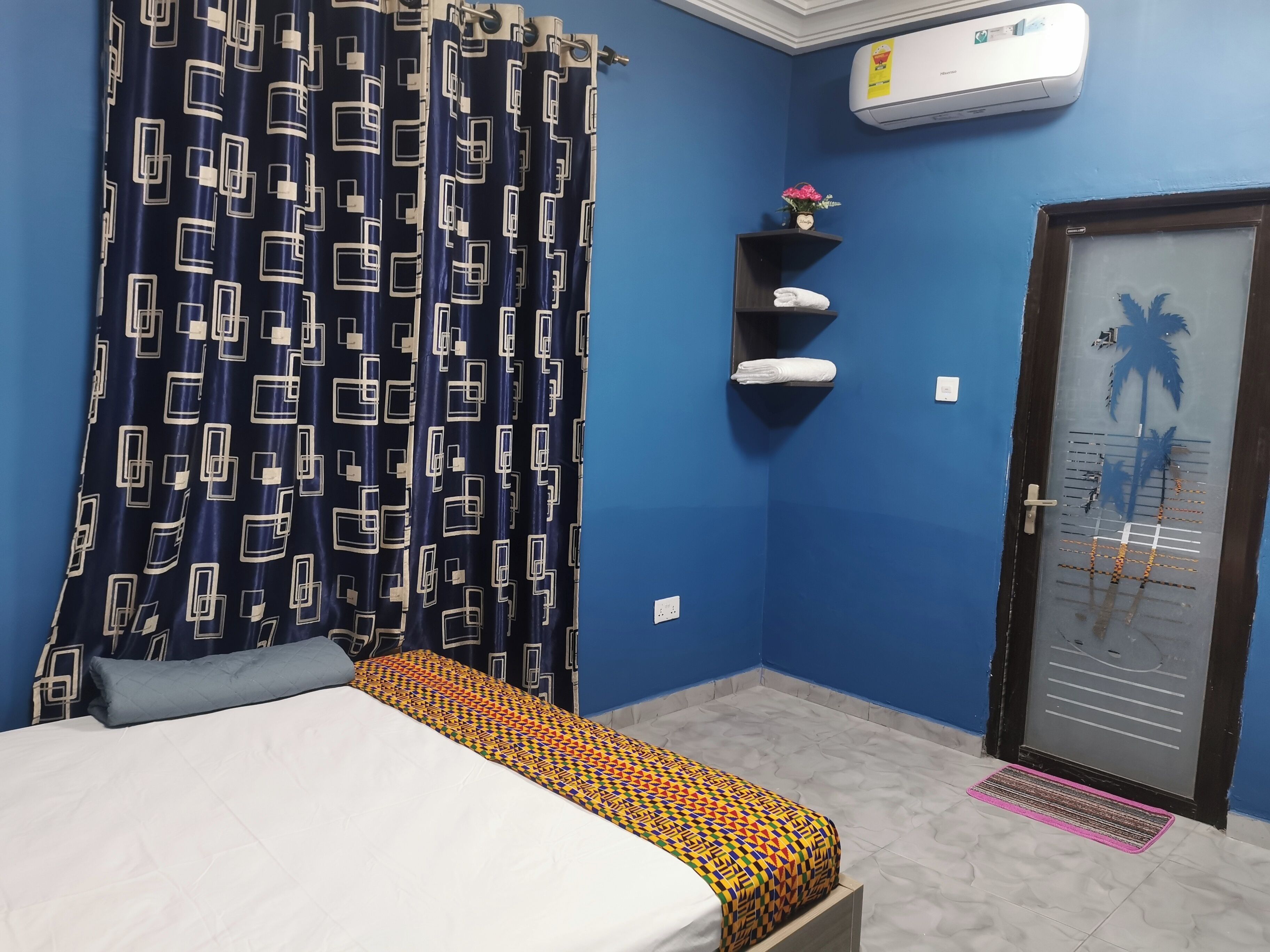 Une chambre avec un lit, un mur bleu, un climatiseur, une porte décorée d’un motif de palmier et une étagère avec des serviettes.