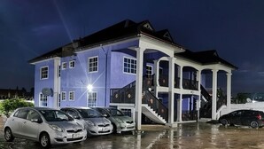 A two-story building with a covered porch and a staircase leading to the upper floor. Several cars are parked in front of the building. - Tropical Oasis in Accra - 2 Bedroom Apartment (Accra)