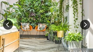 Une chambre d’hôtel avec un parquet en chevrons, un lit en bois, deux fauteuils orange et un mur recouvert de différentes plantes.