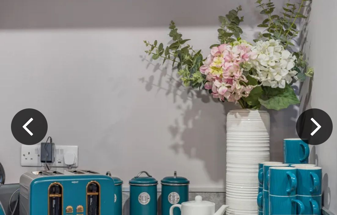 A kitchen counter with a vintage toaster, two teal containers, a white teapot, and a vase with pink and white flowers.