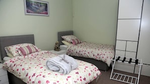 A hotel room with two beds, each with floral bedding and a grey headboard. There is a small bedside table with a lamp and a decorative item. A white wardrobe is against the wall, and a painting hangs above the beds.