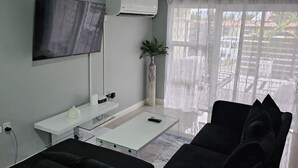 Ett modernt vardagsrum med en platt-TV, luftkonditionering, ett vitt konsolbord, en svart soffa och ett stort fönster med tunna, genomskinliga gardiner.