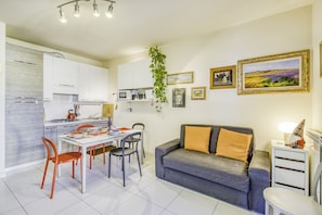 A modern kitchen with white cabinets, a dining table with red chairs, a grey sofa with orange cushions, and a small side table with a lamp. - Appartamento Le Mimose In Lucca, Lucca, Italy (Lucca)