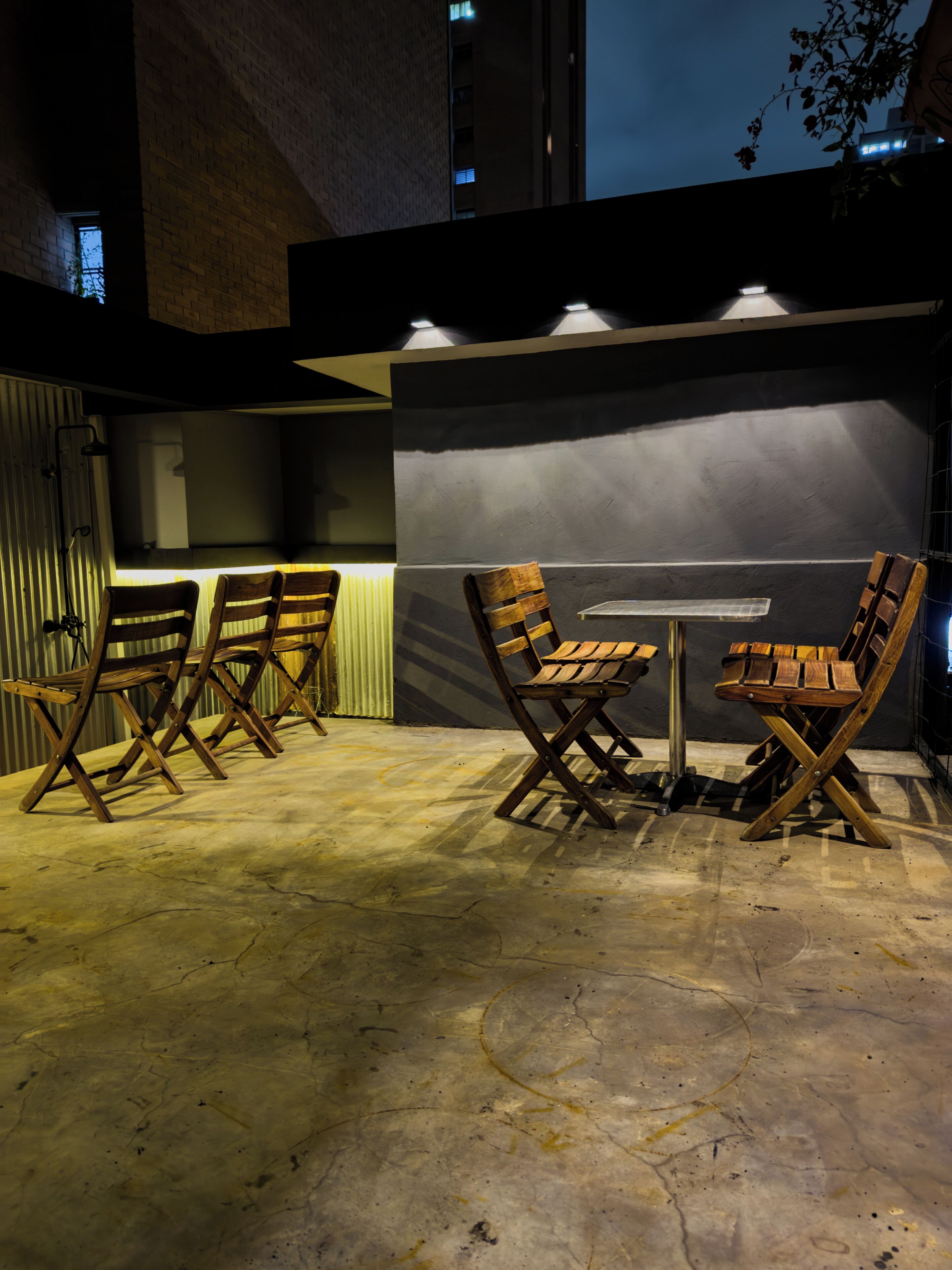 Un espace extérieur aménagé avec des chaises et une table en bois, éclairé par des lampes suspendues, le tout mis en valeur par la façade d’un bâtiment moderne.