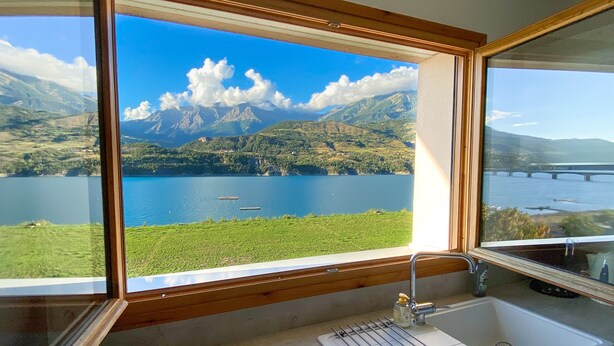 Un baño con un amplio ventanal que ofrece vistas a un lago, montañas y un puente.