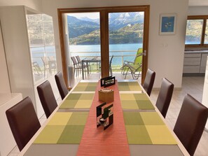 Un espace repas avec une table dressée pour six personnes, des chaises, un buffet et une vue sur un lac et des montagnes, le tout visible à travers de grandes fenêtres.