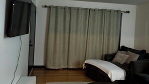 Una sala de estar con un televisor de pantalla plana montado en la pared, un sofá, una mesa de centro y un ventanal con cortinas.