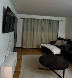 A living room with a flat-screen TV mounted on the wall, a sofa, a coffee table, and a large window with curtains.