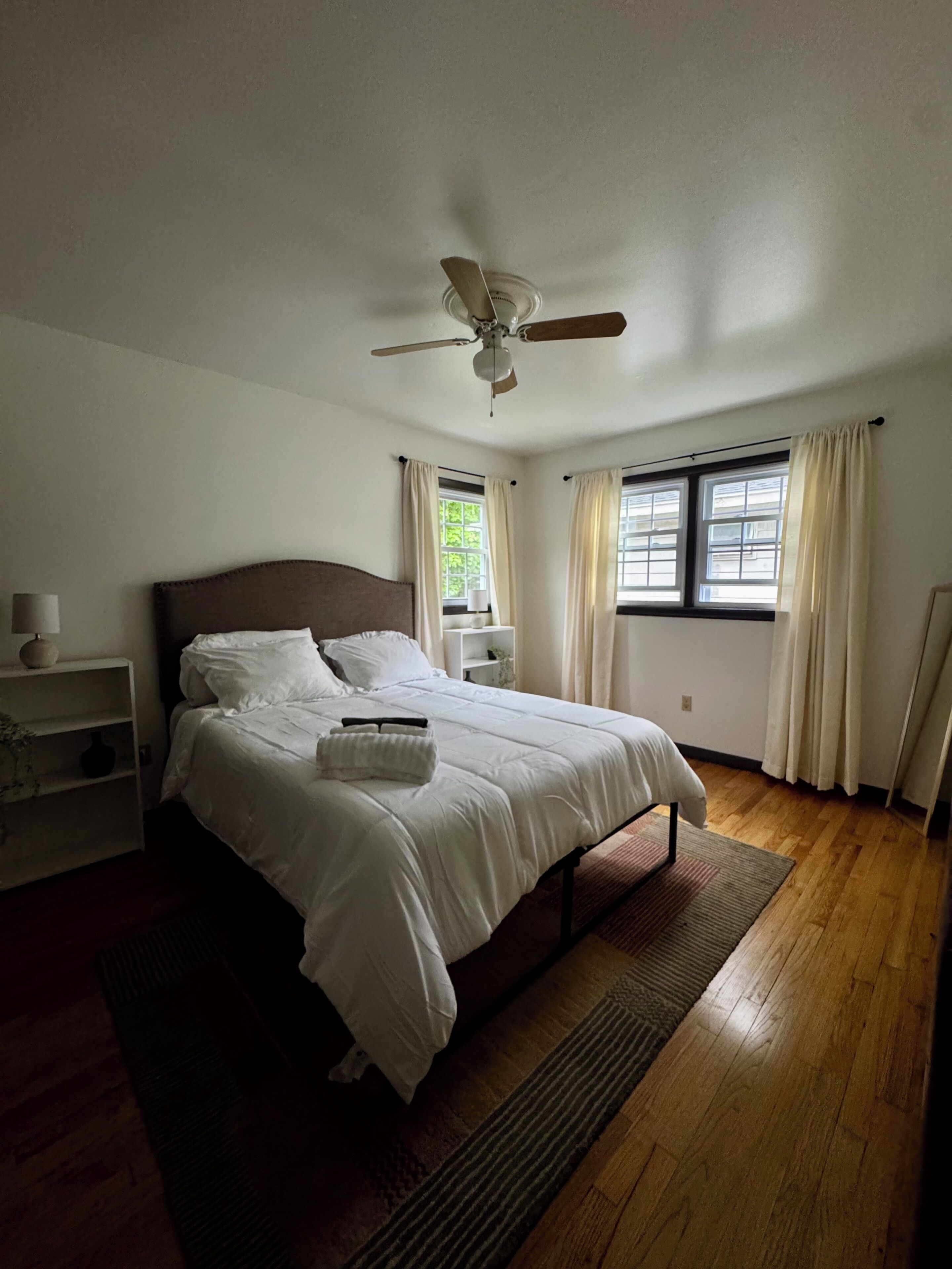 A bedroom with a bed, a nightstand, a ceiling fan, a window with curtains, and a wooden floor.