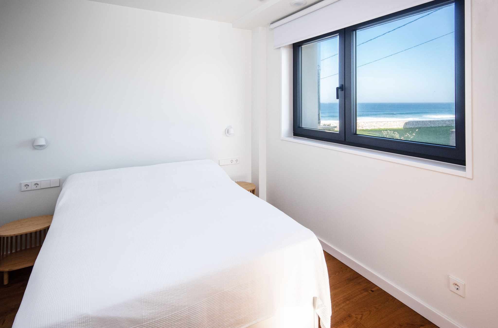 A minimalist bedroom with a single bed, a small wooden stool, a window with a view of the beach, and a white wall.