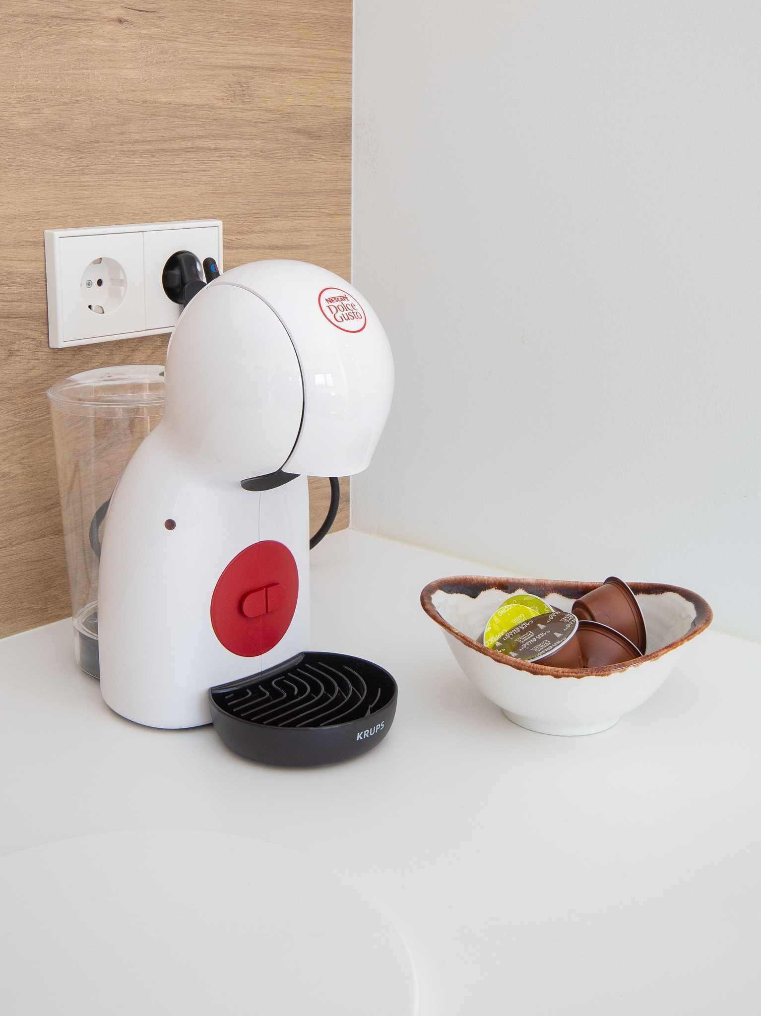 A white coffee machine with a red button, a black remote control, and a bowl with coffee cups on a white countertop.