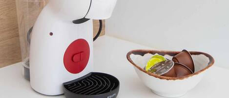A white coffee machine with a red button, a black remote control, and a bowl with coffee cups on a white countertop.