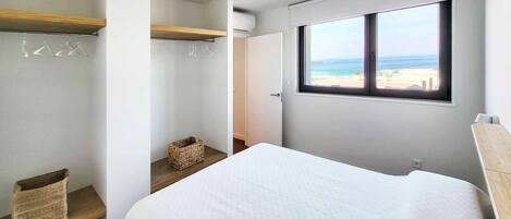 Un dormitorio con cama, ventana con vista a la playa, armario y estante de madera pequeño.
