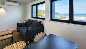 A small room with a sofa, a table, and two chairs. There are two windows with a view of the beach and the sea.