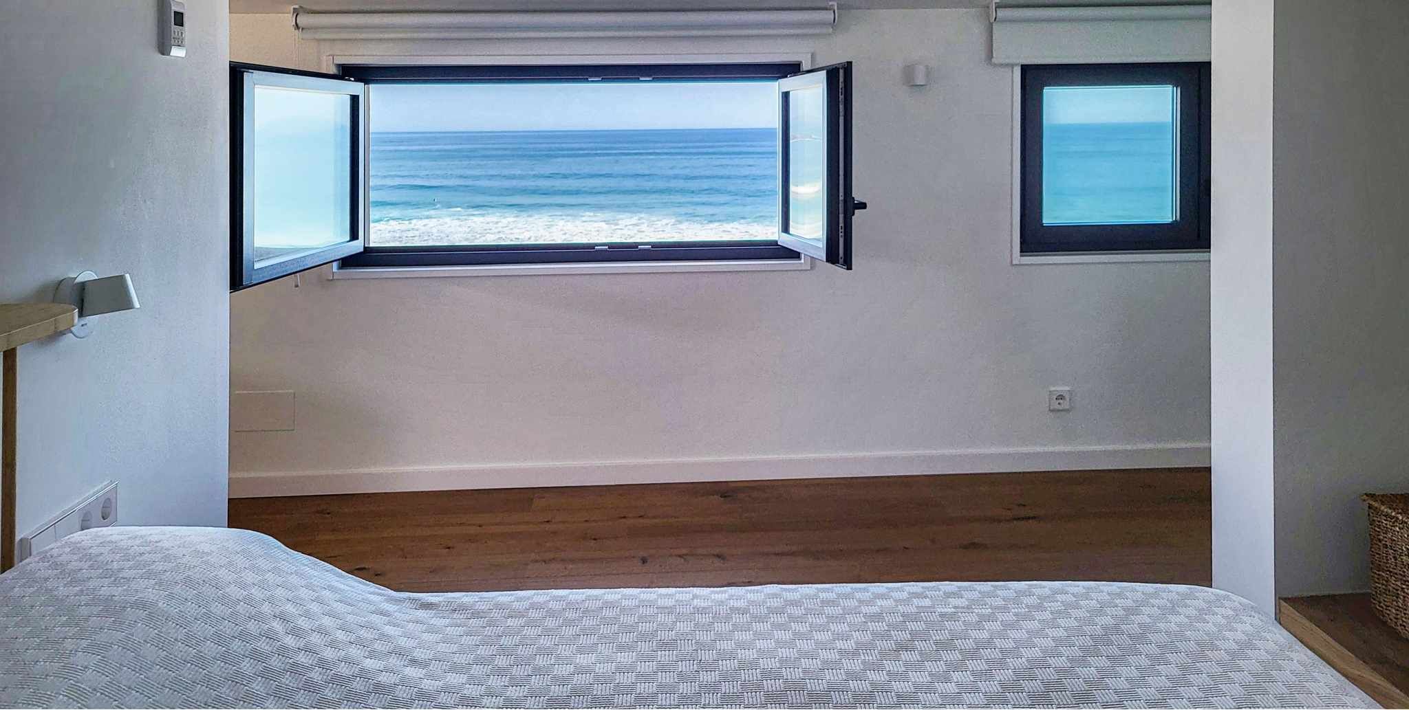 A bedroom with a large window overlooking the sea, a bed, and a small window.