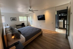 A modern bedroom with a bed, a desk, a chair, a ceiling fan, a TV, and a door leading to a kitchen.